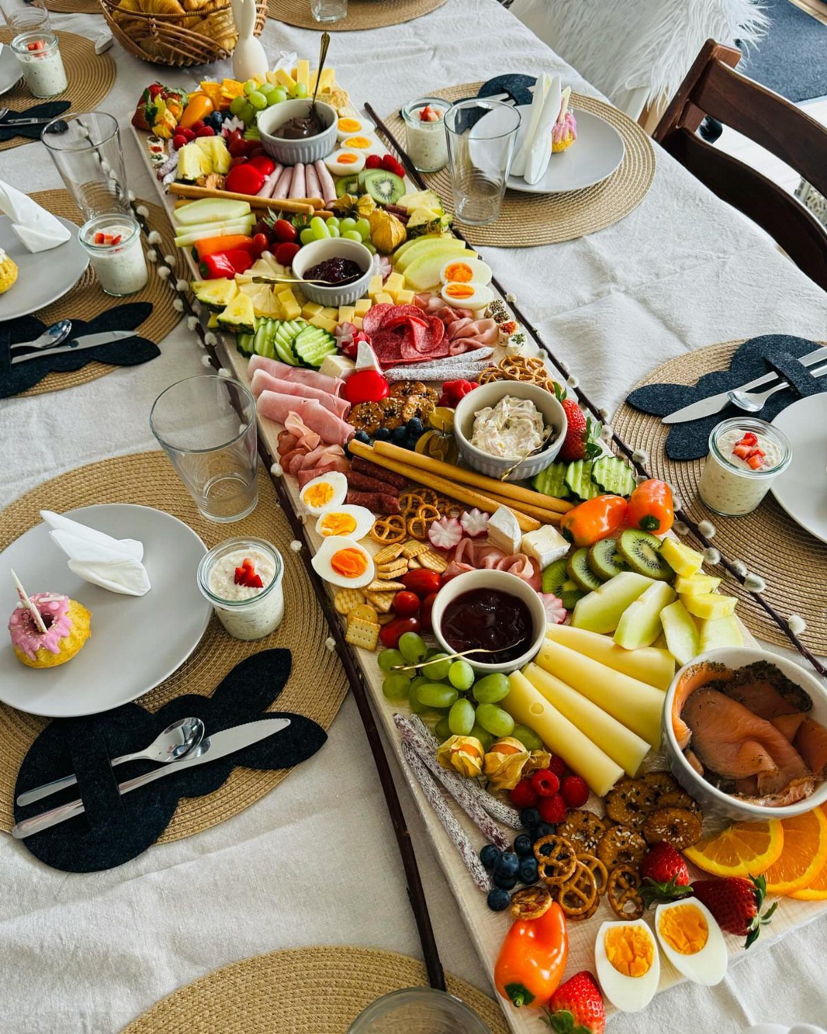 Long charcuterie platter with meats, cheeses, fruits, and eggs on a white table.