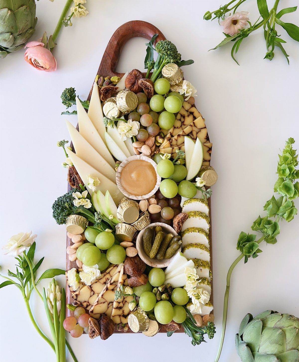 Wooden charcuterie board with green grapes, cheeses, and chocolates surrounded by spring flowers.