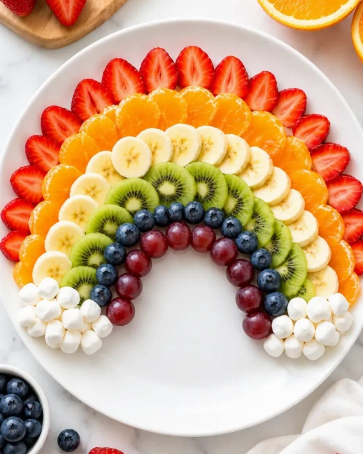 Rainbow fruit arrangement with strawberries, oranges, bananas, kiwi, grapes, and marshmallows on plate