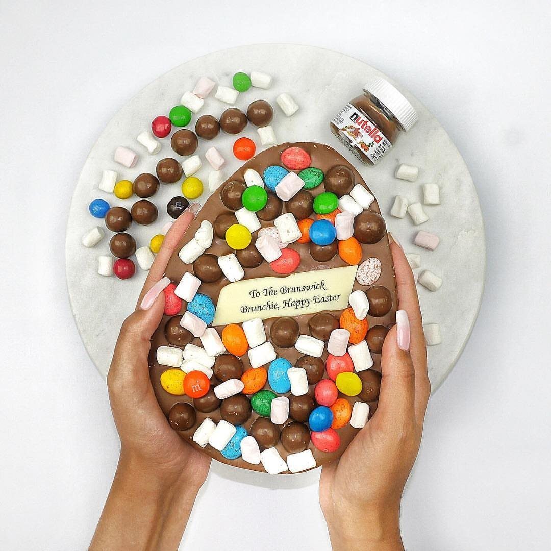 Large chocolate Easter egg decorated with colorful candies and marshmallows held by hands.