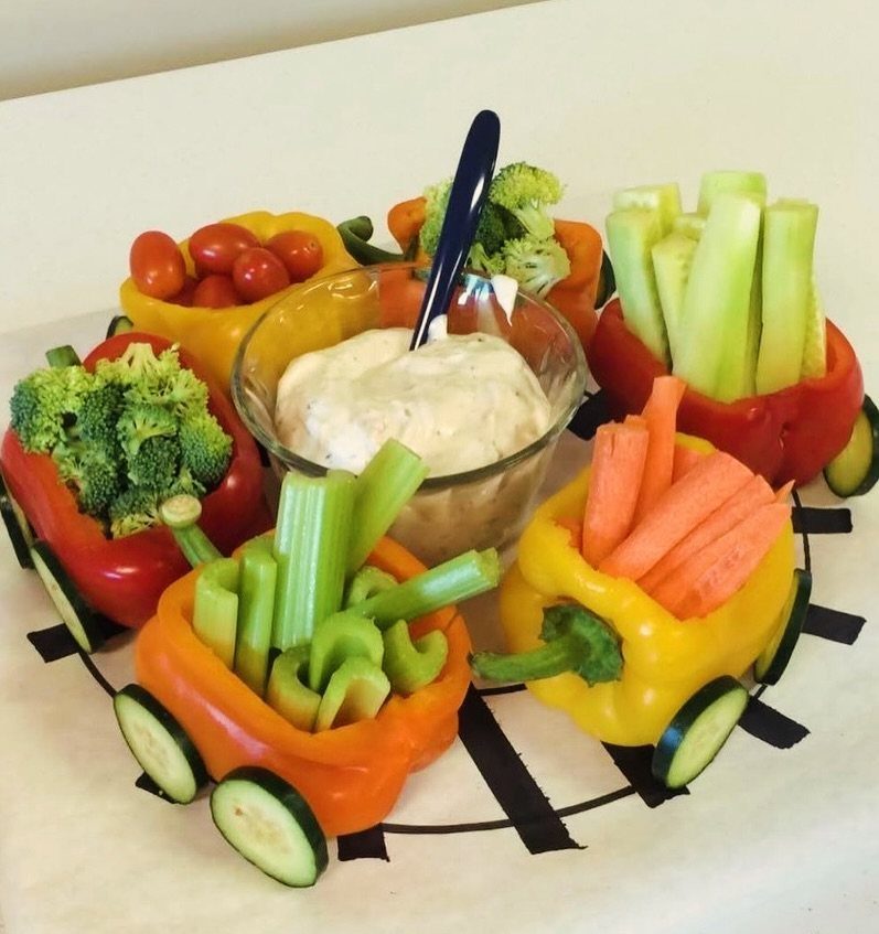 Bell pepper cars filled with fresh vegetables surrounding a central glass dip bowl.