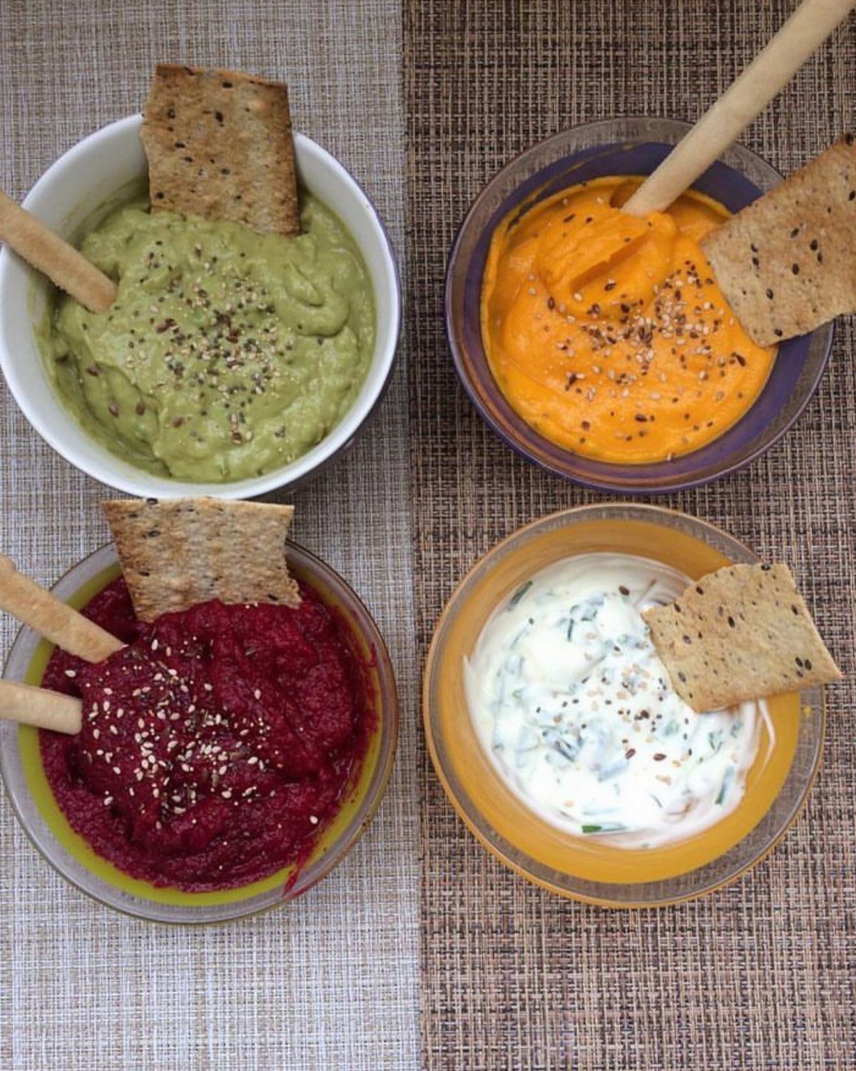 Four colorful homemade dips in bowls served with crackers and breadsticks.