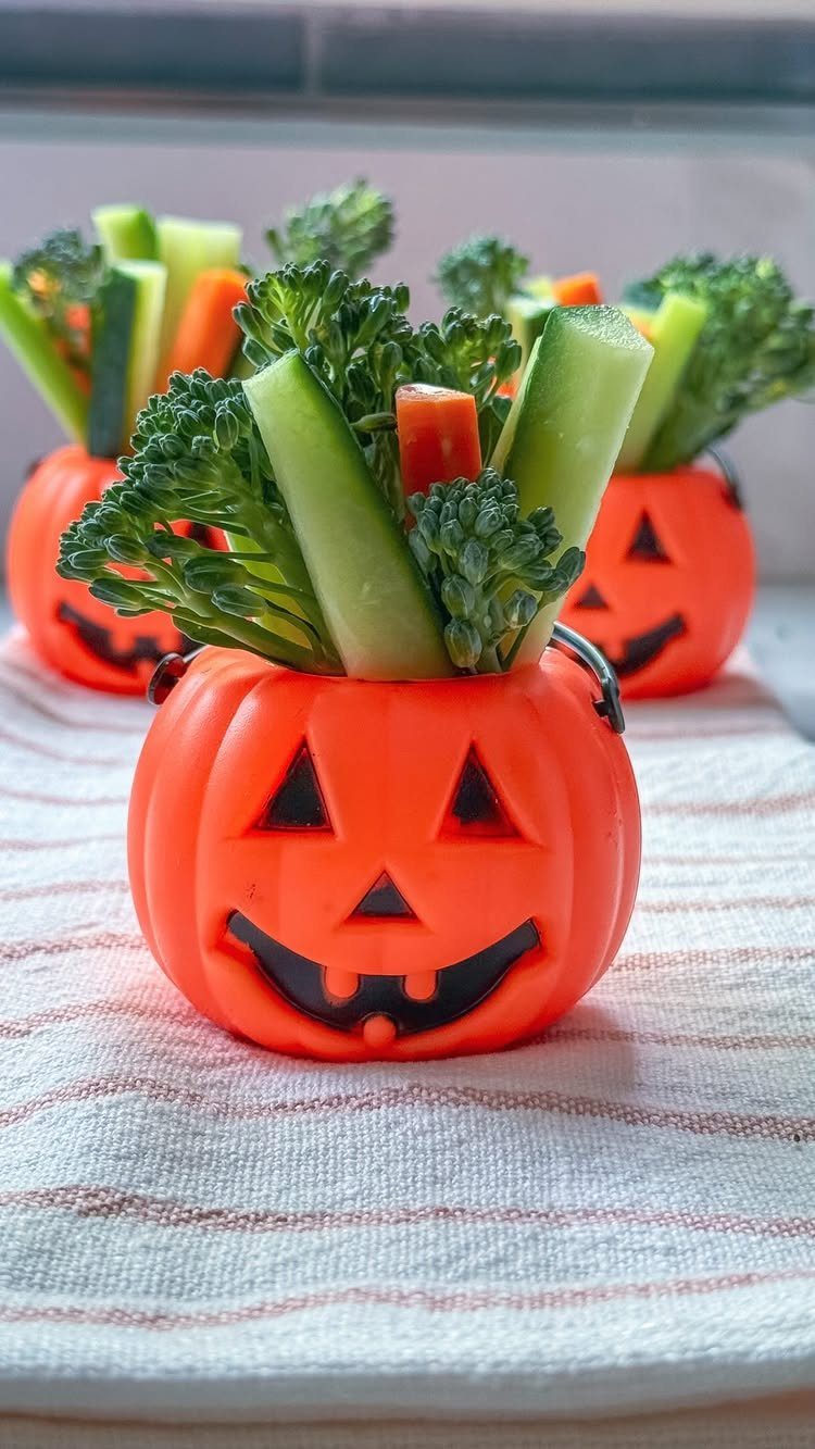 Orange jack-o'-lantern buckets filled with broccoli, celery, and carrot sticks on striped cloth.