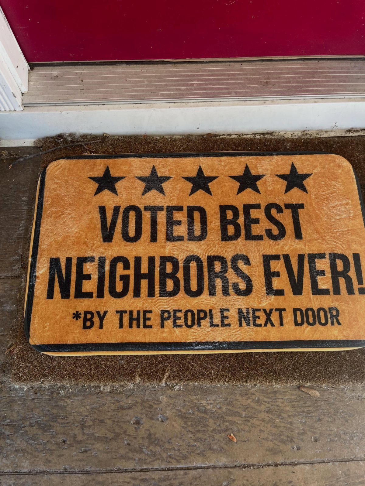 Brown coir doormat with funny text about best neighbors on a wooden porch.