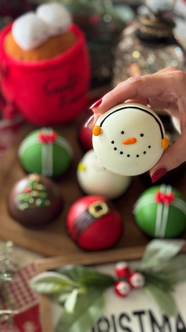 Hand holding a snowman-shaped hot chocolate bomb among other festive chocolate spheres.