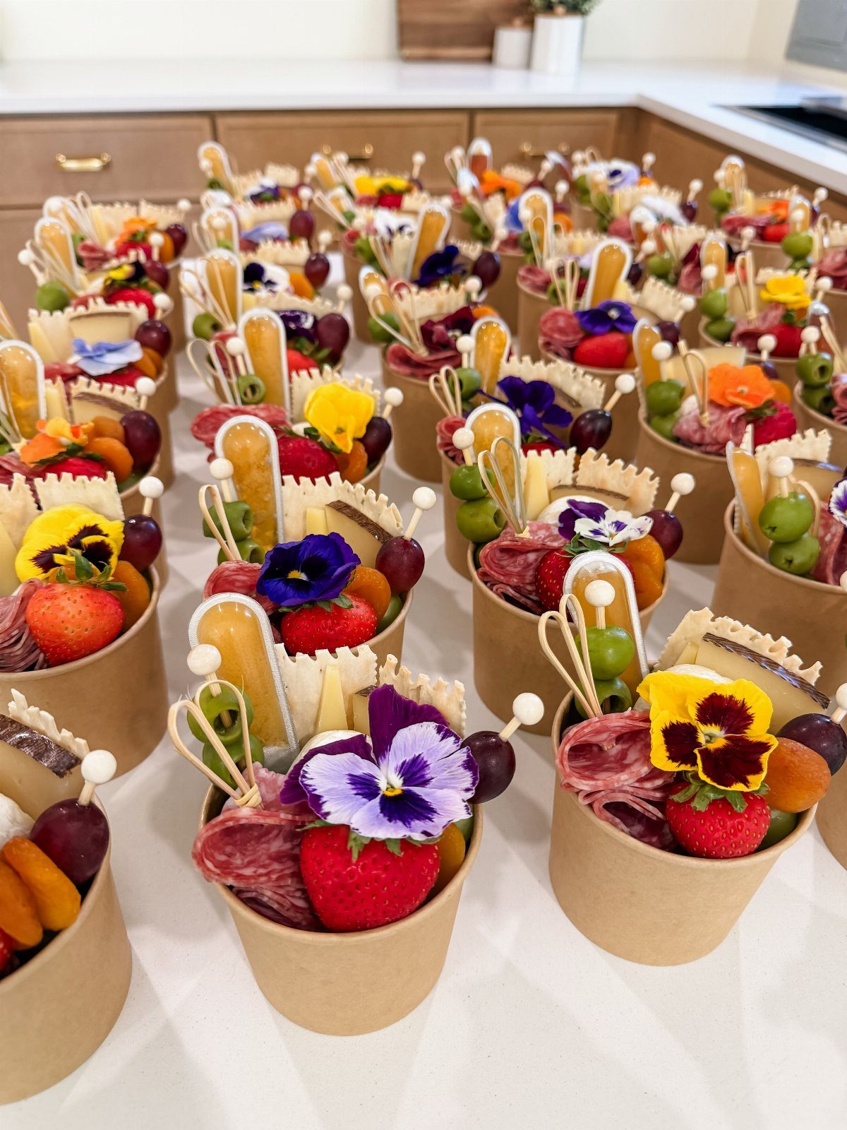 Charcuterie cups filled with meats, cheeses, fruits, and edible flowers on white counter