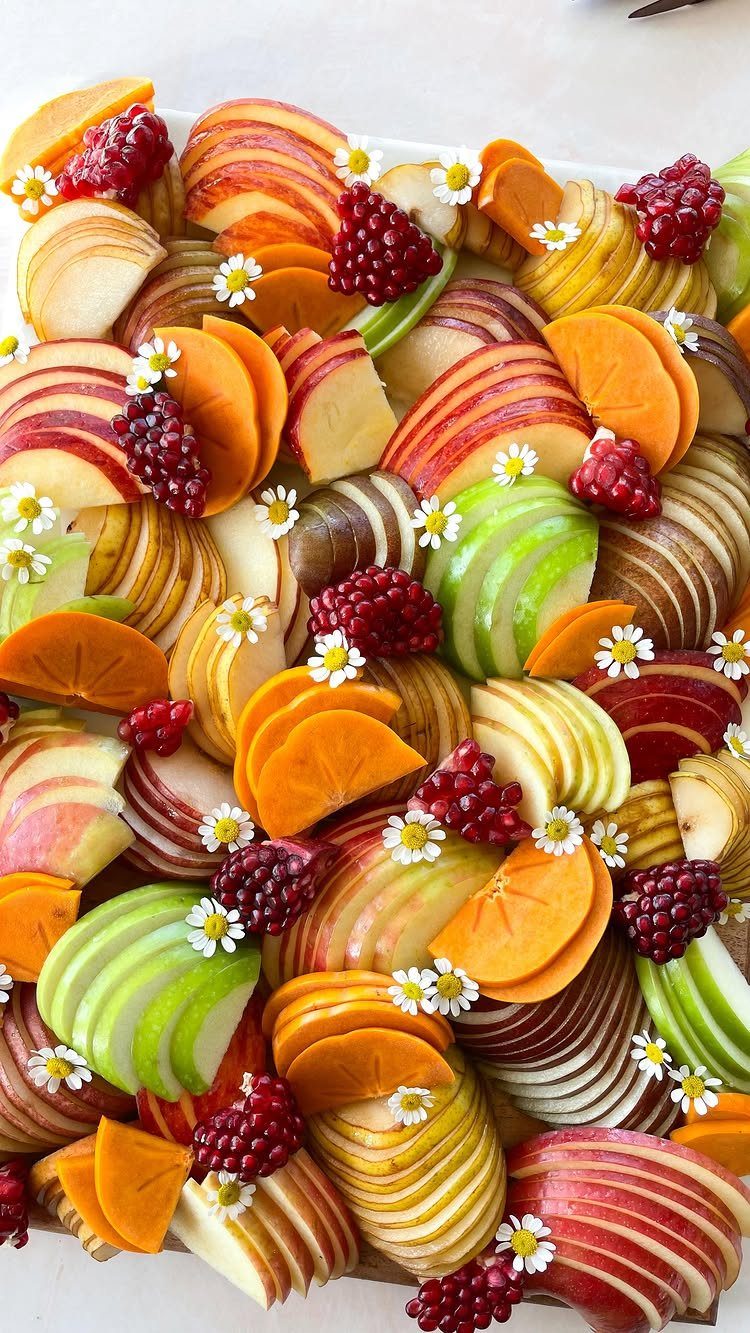 Sliced apples, persimmons, and pomegranate seeds arranged on a platter with white daisies.