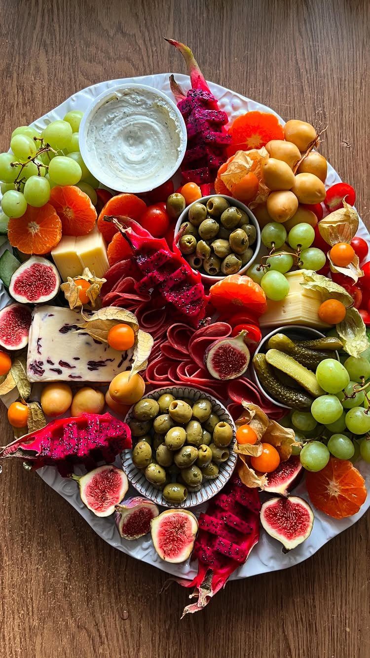 Charcuterie platter featuring bowls of green olives, gherkin pickles, grapes, and sliced figs.