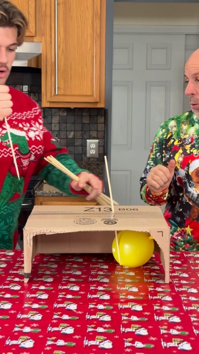 Two men in Christmas sweaters playing a balloon popping game with wooden skewers