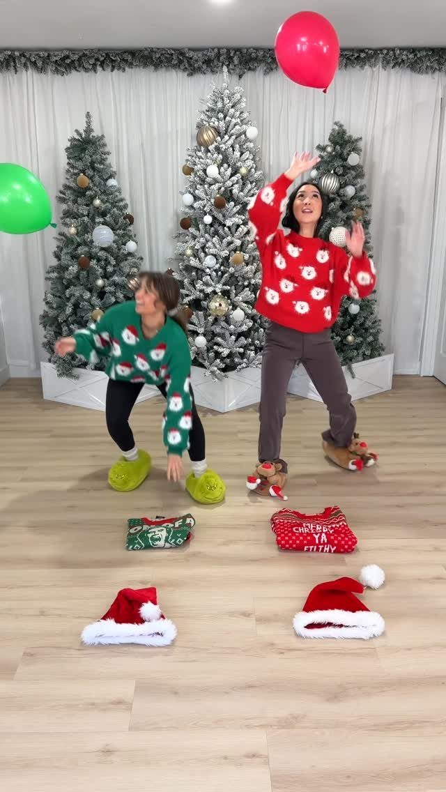 Two women in Christmas sweaters playing a balloon game near decorated holiday trees.