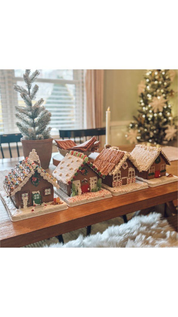 Four decorated gingerbread houses lined up on a wooden table near a Christmas tree.