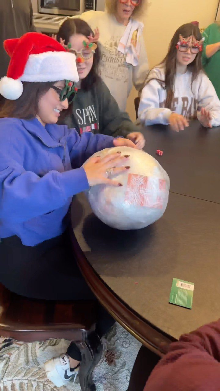 Woman in Santa hat unwrapping a large plastic-wrapped prize ball at a table.