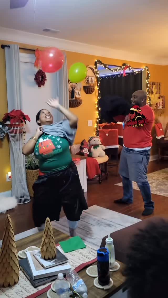 Woman in Christmas sweater laughing while playing with red and green balloons indoors.