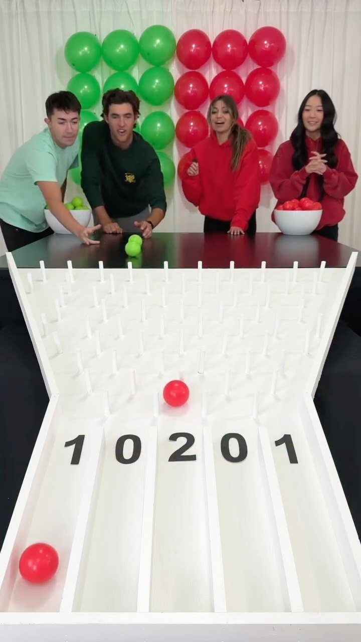 Four people playing a Plinko game with red balls and balloon decorations.