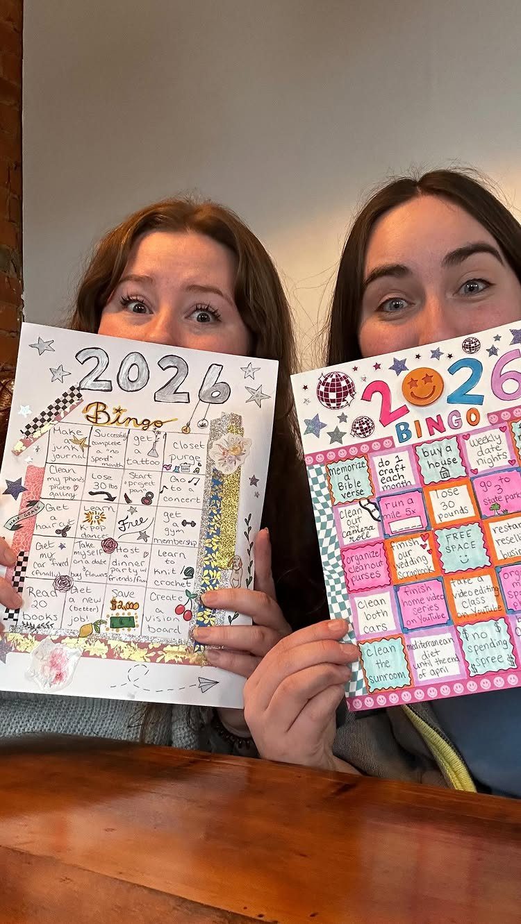 Two women holding colorful handmade 2026 bingo cards for a New Year's game.