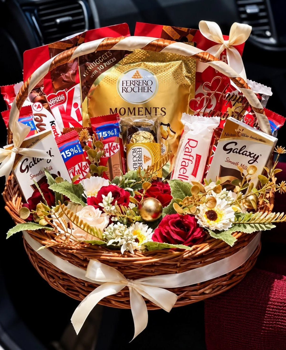 Wicker gift basket filled with assorted chocolates and red and white artificial flowers.