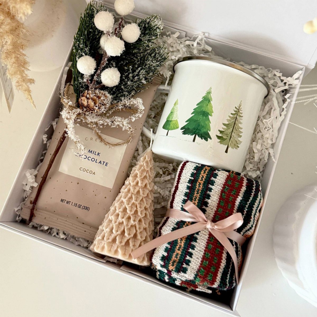 Hot cocoa gift box with chocolate bar, tree-patterned mug, and cozy knitted accessories.