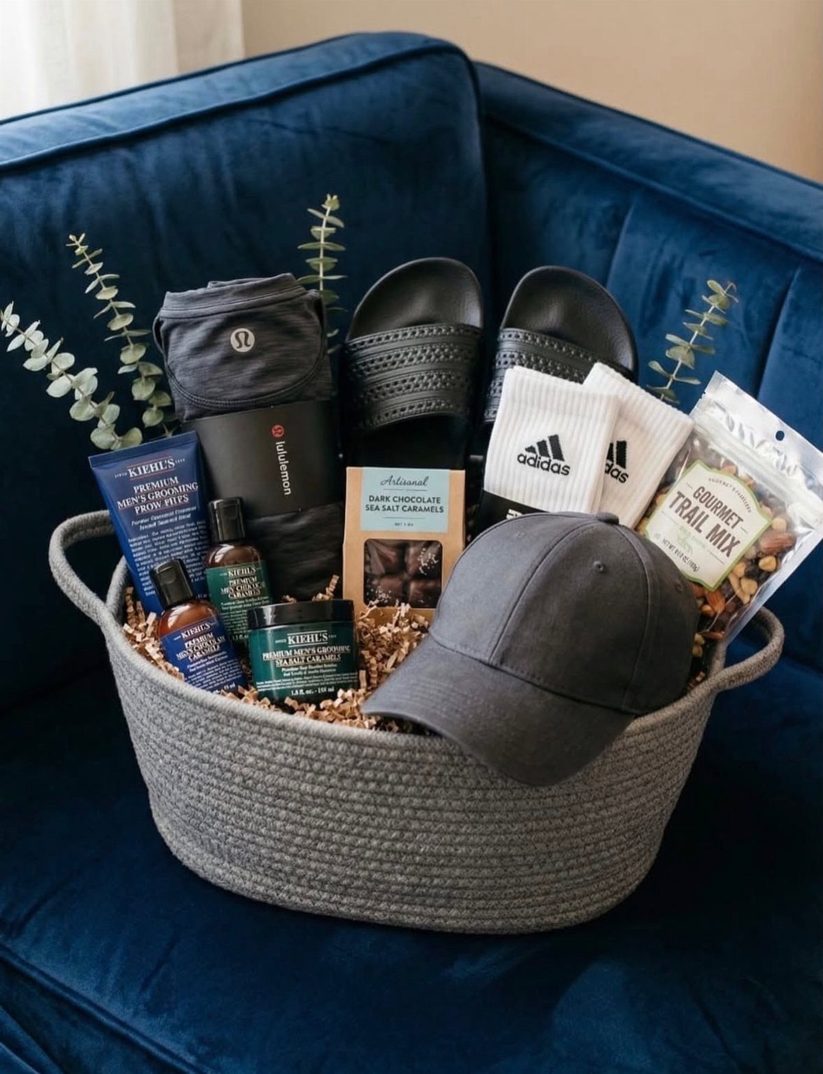 Grey woven gift basket containing athletic apparel, grooming products, and snacks on blue sofa.