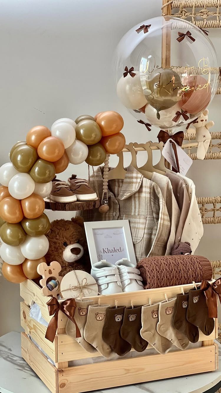 Baby gift basket in a wooden crate with teddy bear, clothes, and balloons.