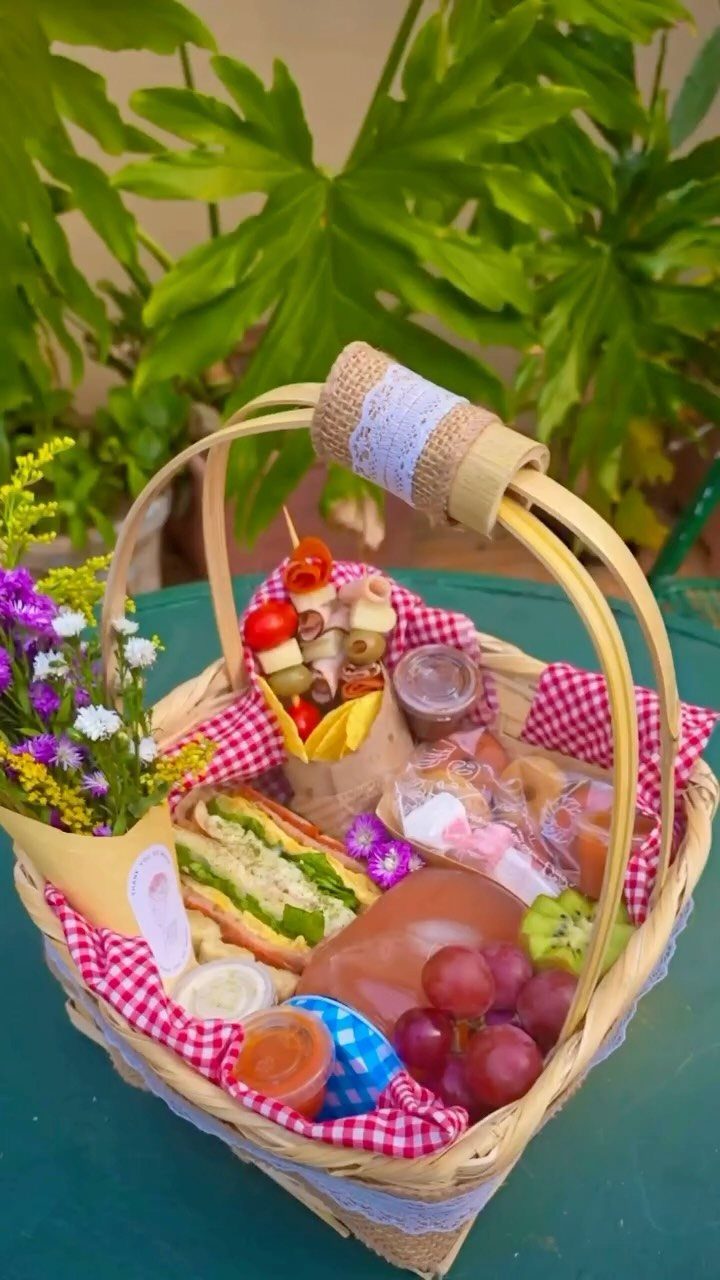 Wicker picnic basket filled with sandwich, grapes, meats, and fresh flowers outdoors.