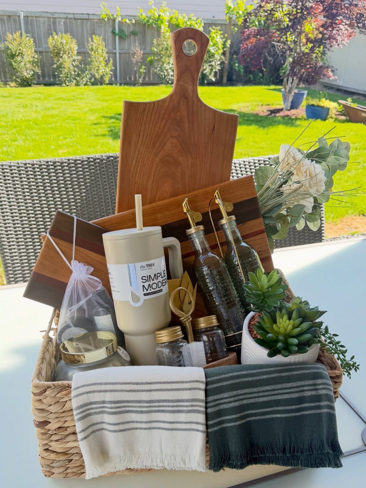 Woven gift basket with wooden board, tumbler, bottles, and towels on outdoor table