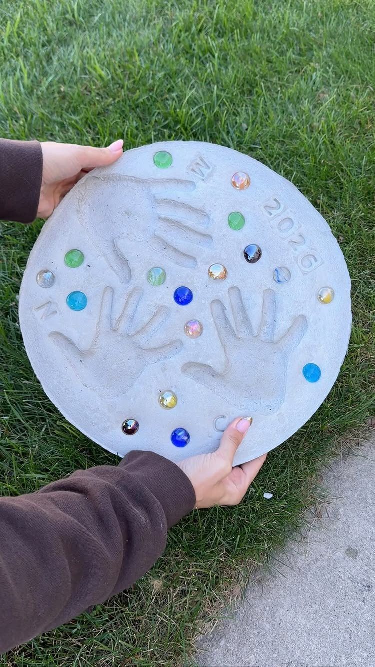 Circular concrete garden stone with handprints and colorful gems held over green grass