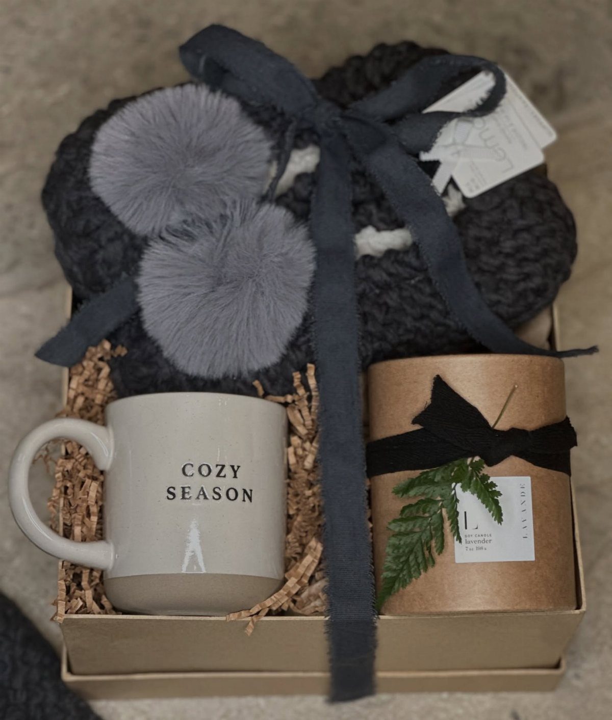 Gift box with a grey knitted blanket, cozy season mug, and wrapped treat.