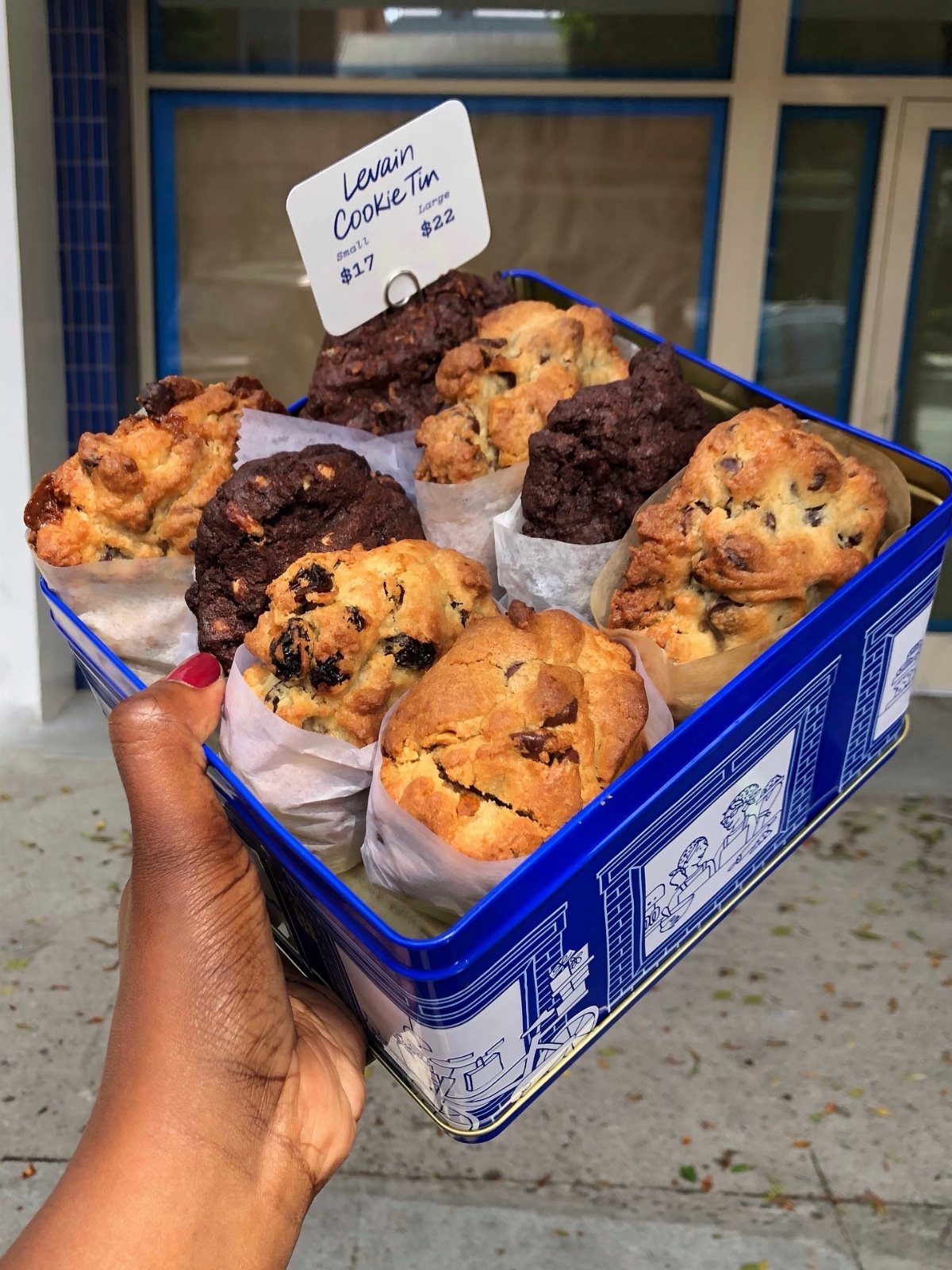 Blue metal tin filled with large chocolate chip cookies held by a hand.