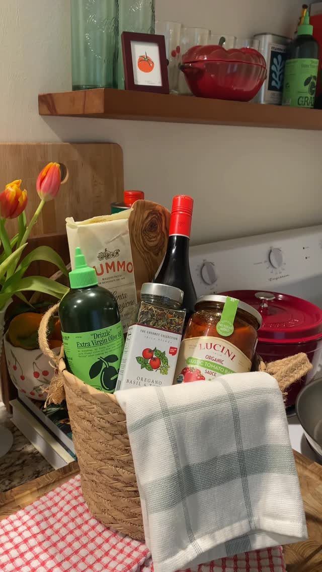 Woven gift basket with pasta, sauce, olive oil, and wine on checkered cloth.