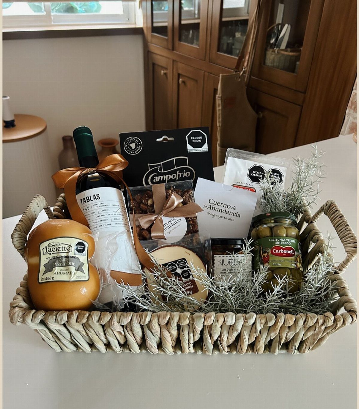 Woven gift basket with wine, cheese, olives, and gourmet snacks on a table.