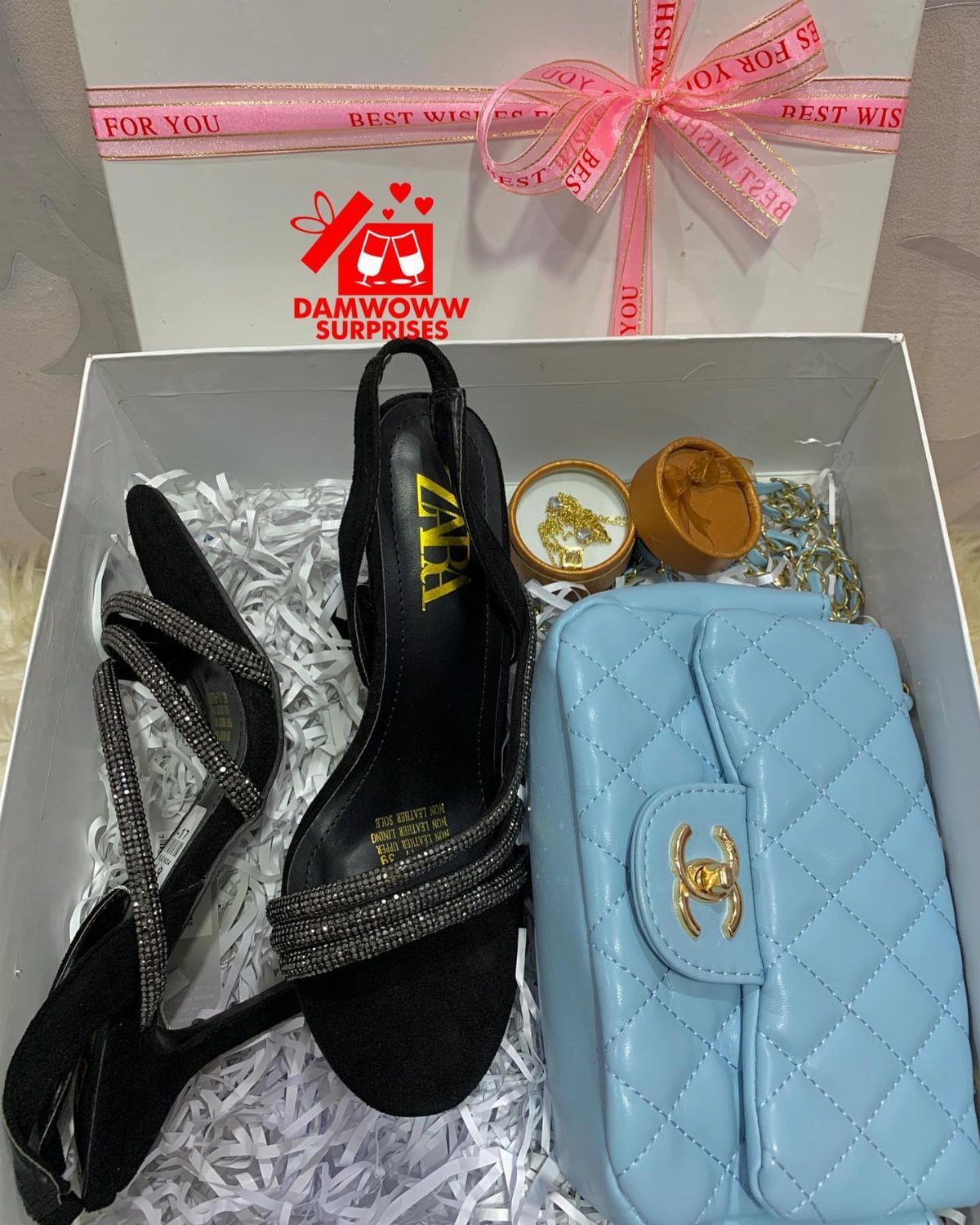 Gift box containing black high heels, a light blue quilted purse, and small jars.
