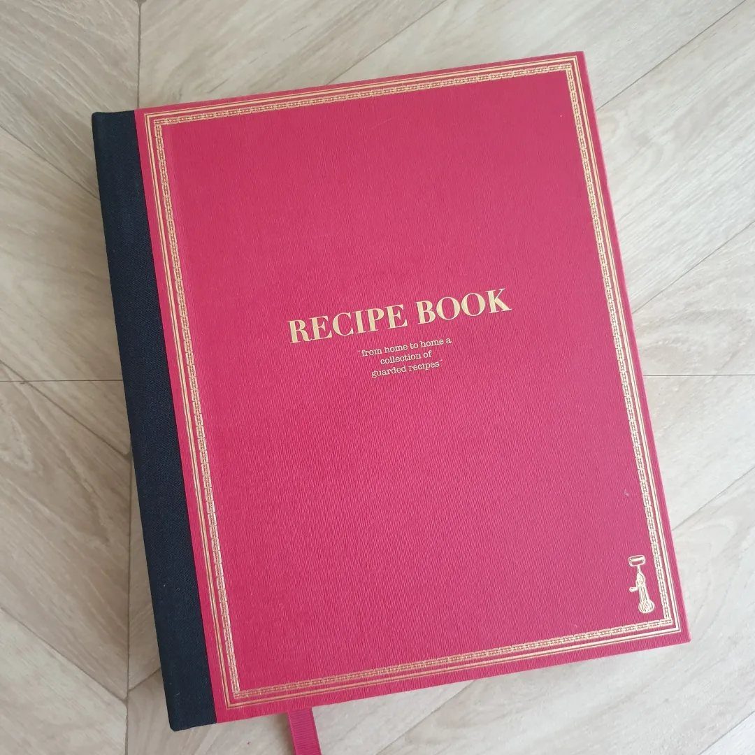 Red recipe book with gold lettering resting on a light wooden floor.