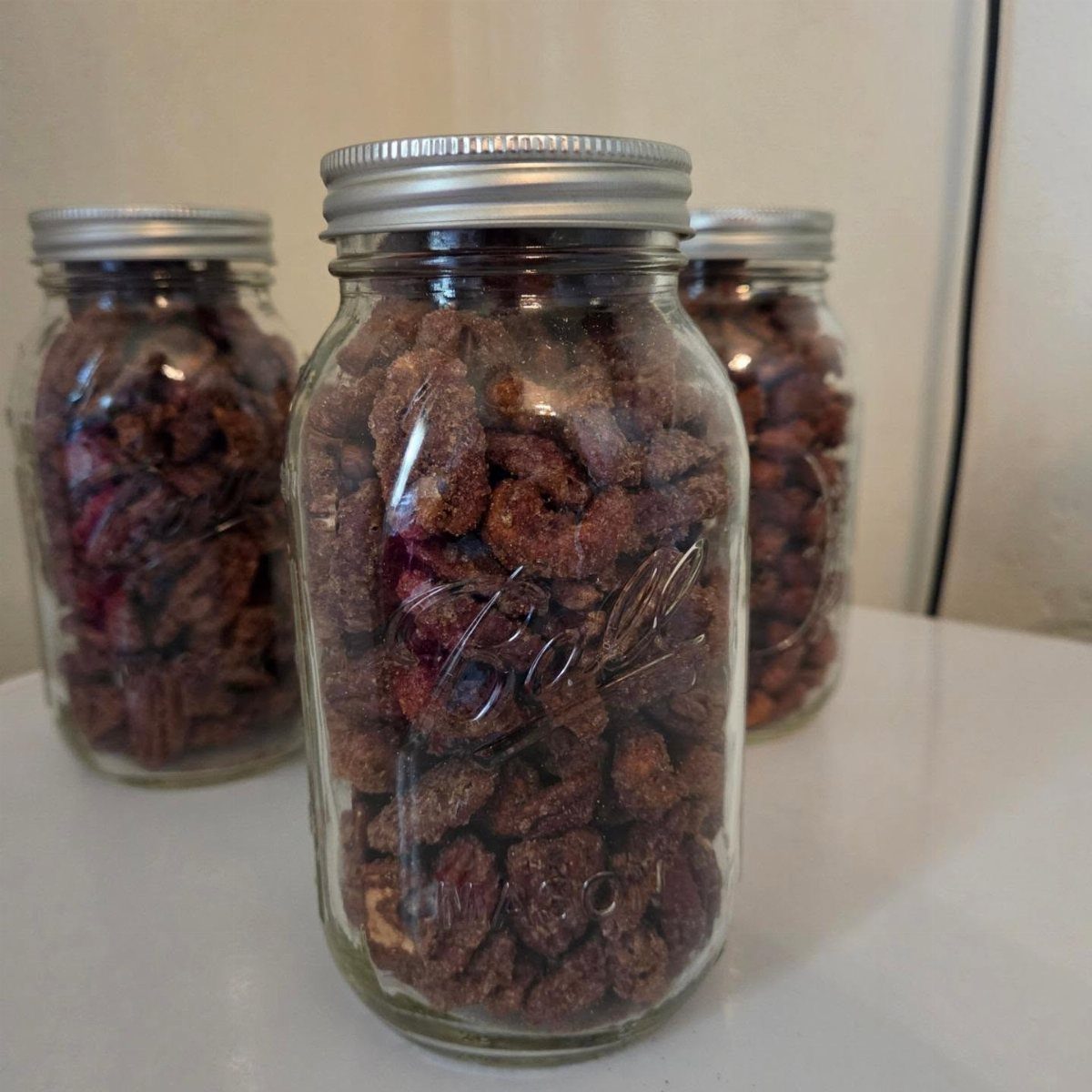 Three mason jars filled with glazed candied pecans on a white surface.