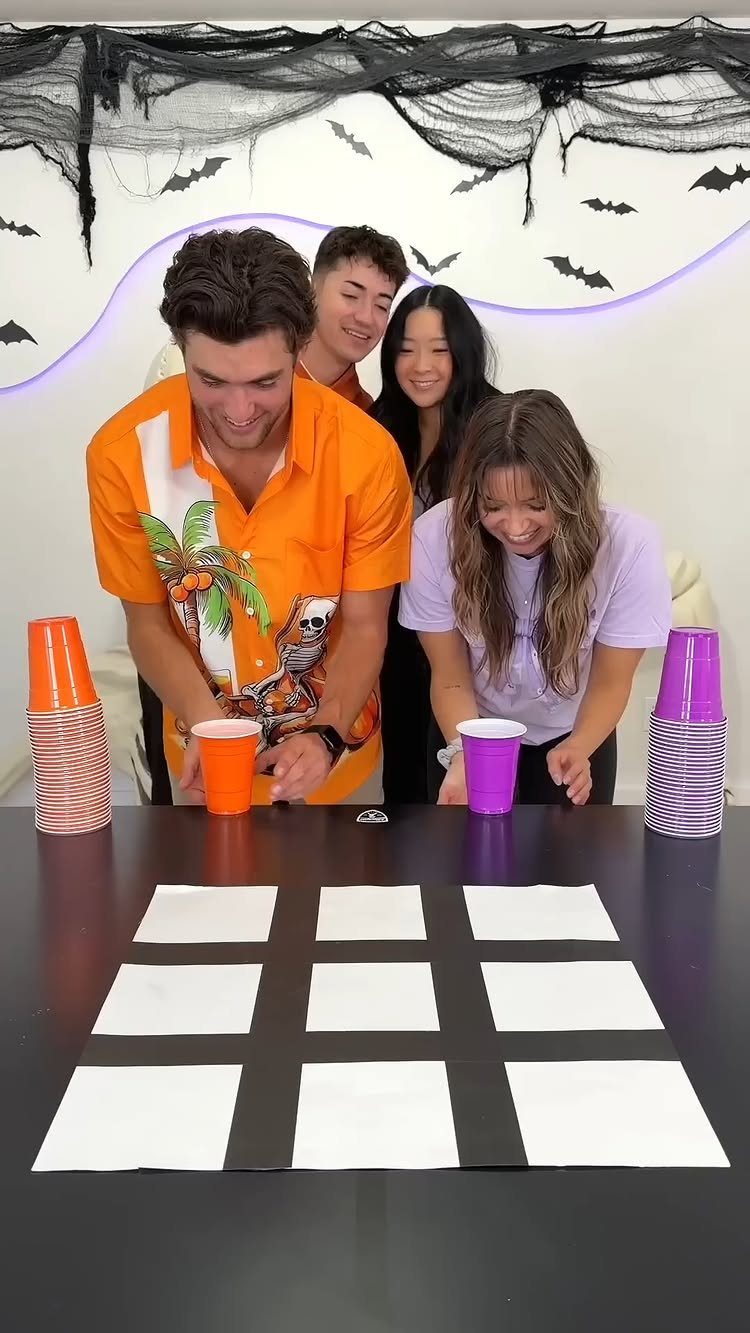 Young adults playing a cup-based game on a grid table with Halloween decorations.