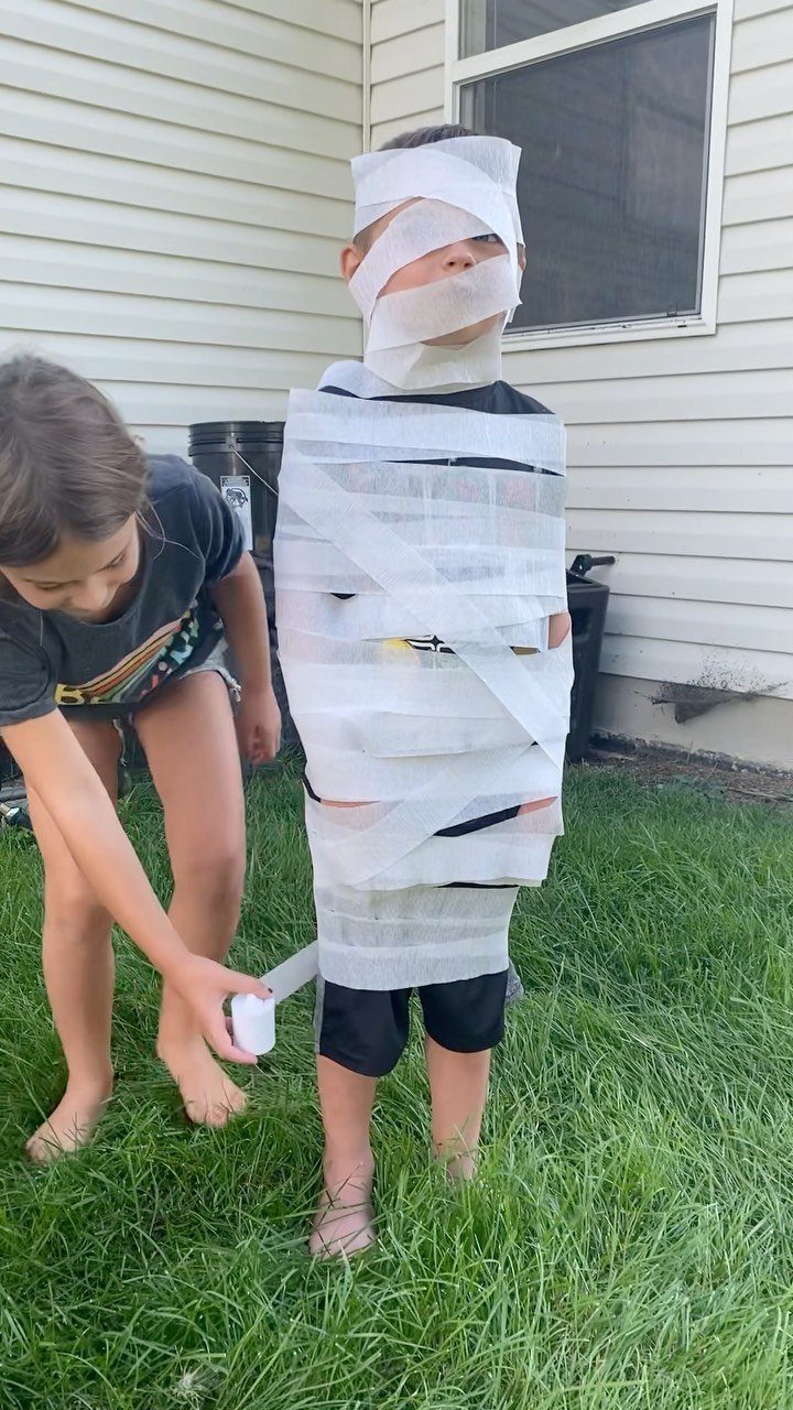 Girl wrapping a boy in toilet paper to look like a mummy outdoors.