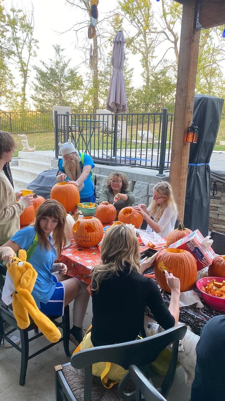 People sitting around an outdoor table carving faces into orange pumpkins for Halloween.