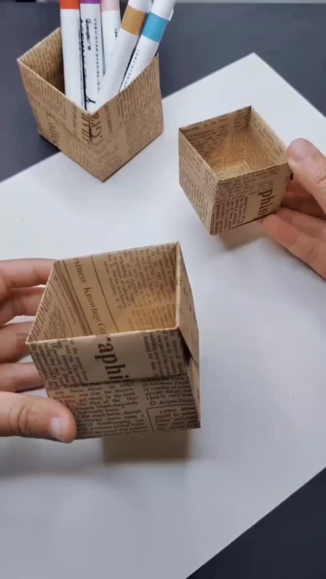 Small brown newspaper-patterned paper boxes, one holding colorful markers on white surface.