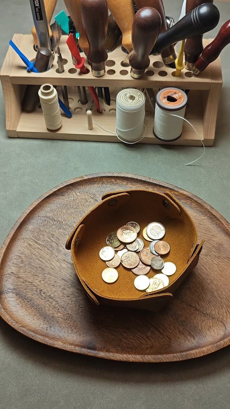 Brown leather catchall tray with coins on a wooden platter and leatherworking tools.
