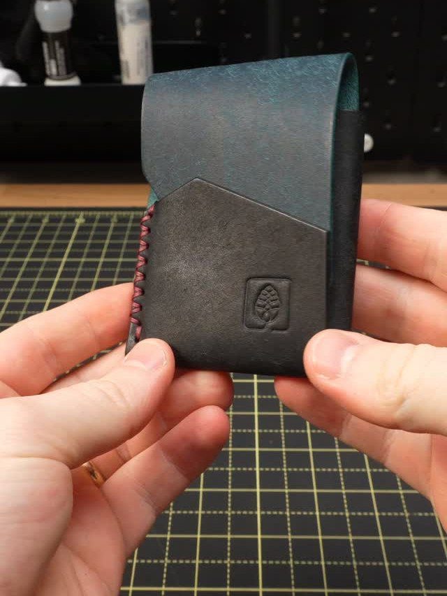 Handmade leather wallet with blue-green and black panels held over a cutting mat.