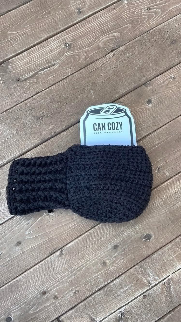 Black crocheted can cozy holding a white beverage can on wooden decking.
