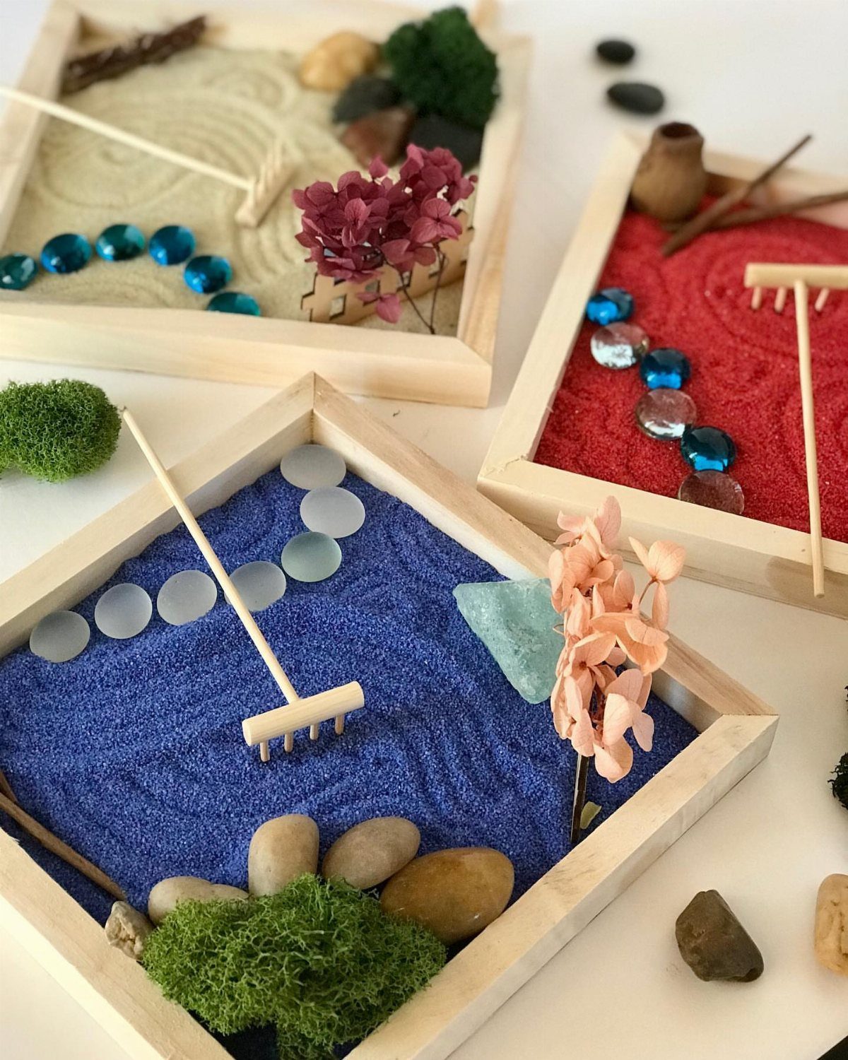 Three miniature zen gardens with colored sand, rakes, stones, and moss in wooden trays.