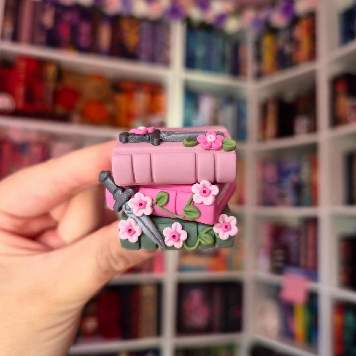 Miniature clay book stack with pink flowers and swords held in a hand.
