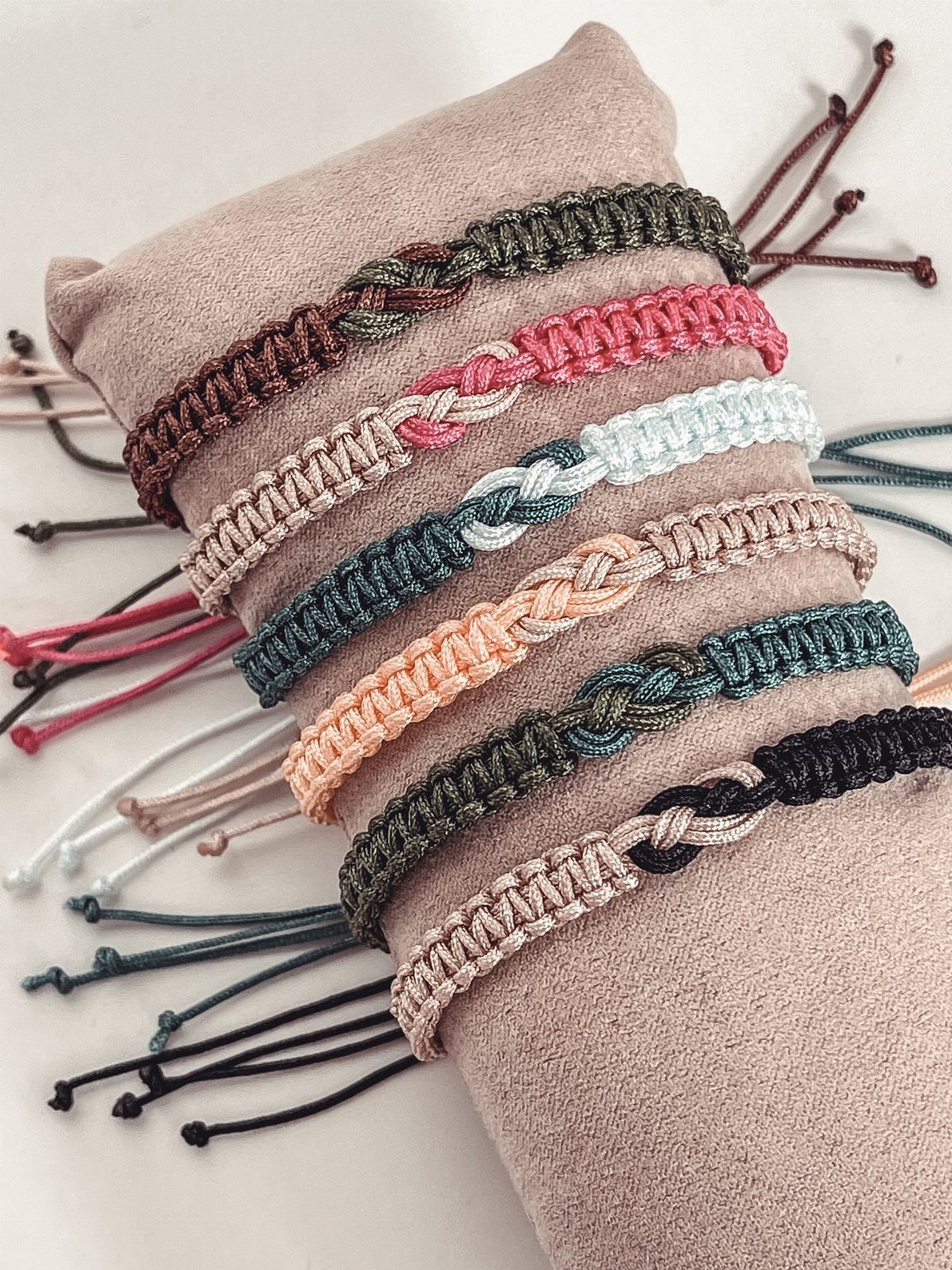 Woven cord bracelets in multiple colors displayed on a tan fabric cushion.