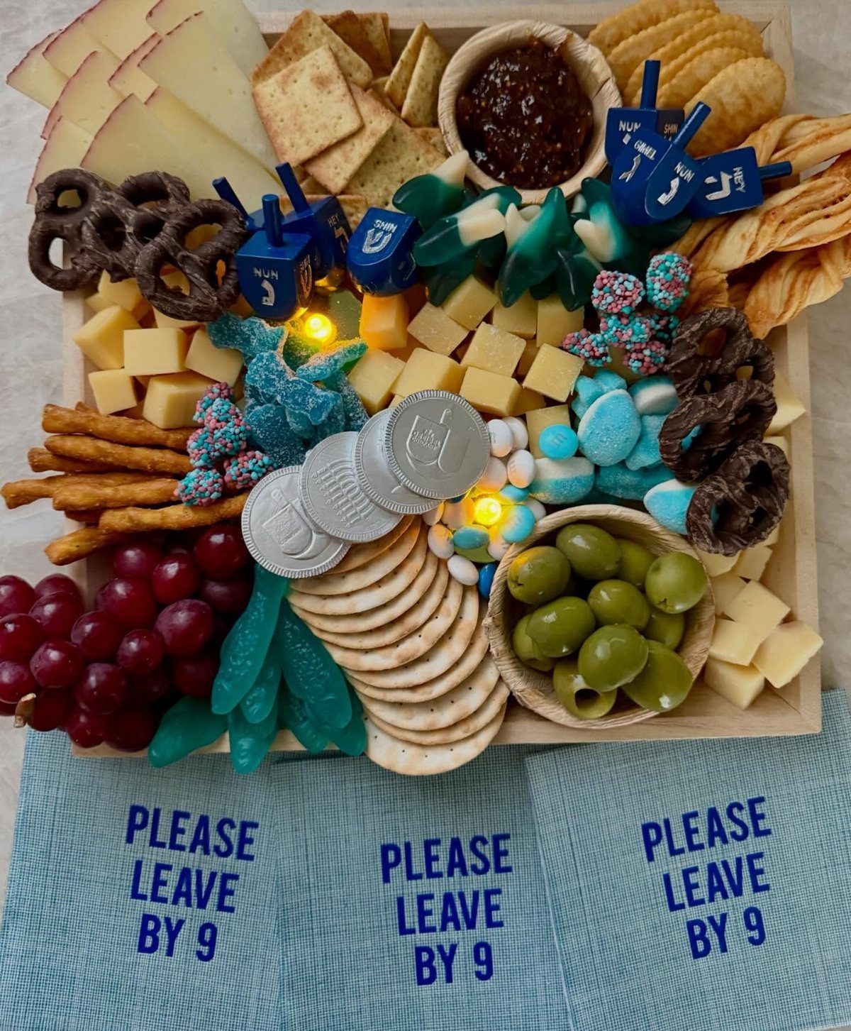 Charcuterie board with cheese, crackers, blue candies, silver coins, and blue dreidels.