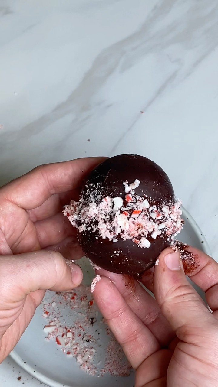 Chocolate sphere decorated with crushed peppermint candies held by two hands.