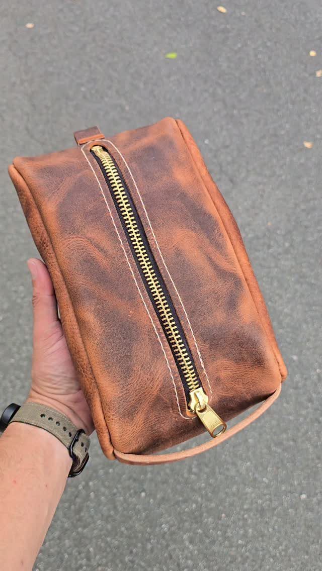 Brown leather dopp kit travel bag with gold zipper held in a hand.