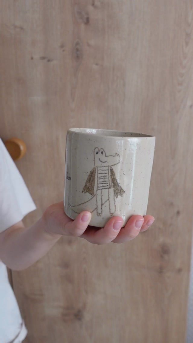 Beige ceramic mug with a child's drawing of a crocodile held in a hand.