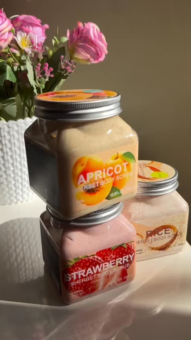 Three square mason jars of fruit-scented body scrubs next to pink roses.