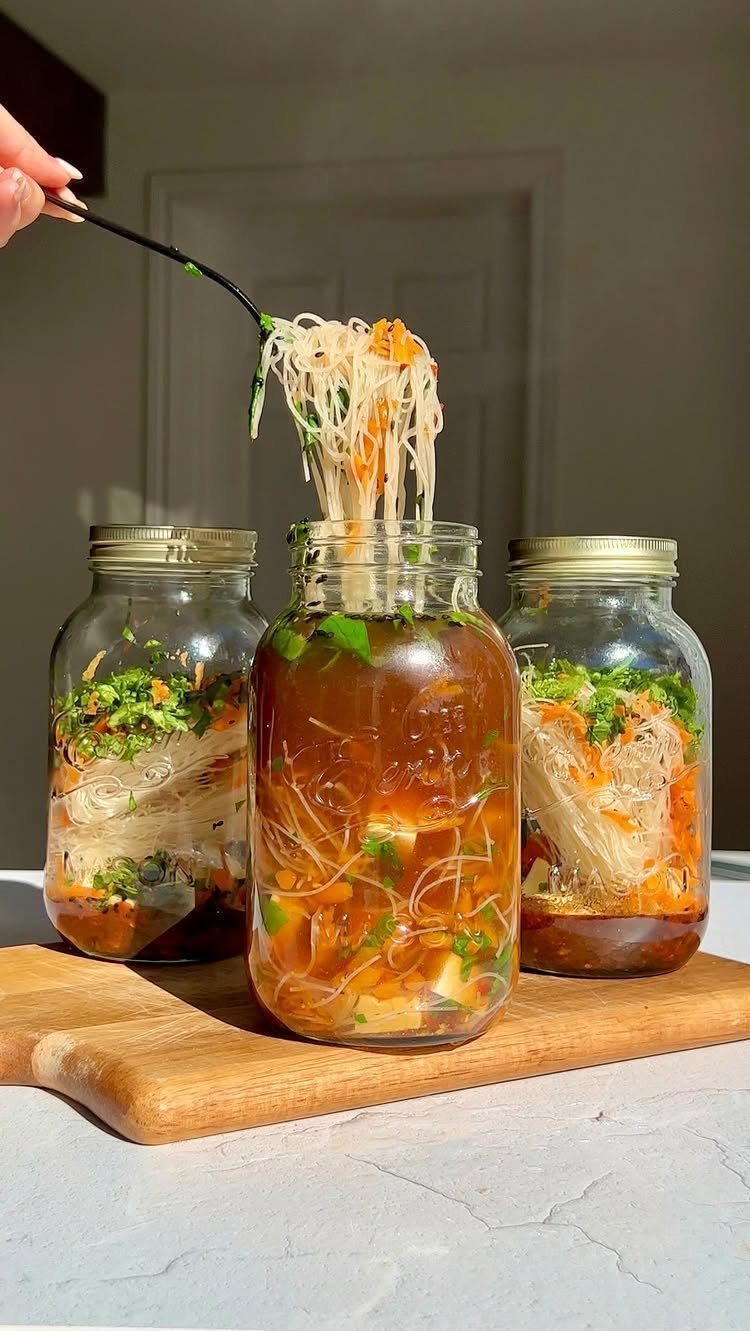 Three mason jars filled with layered noodle soup on a wooden board.