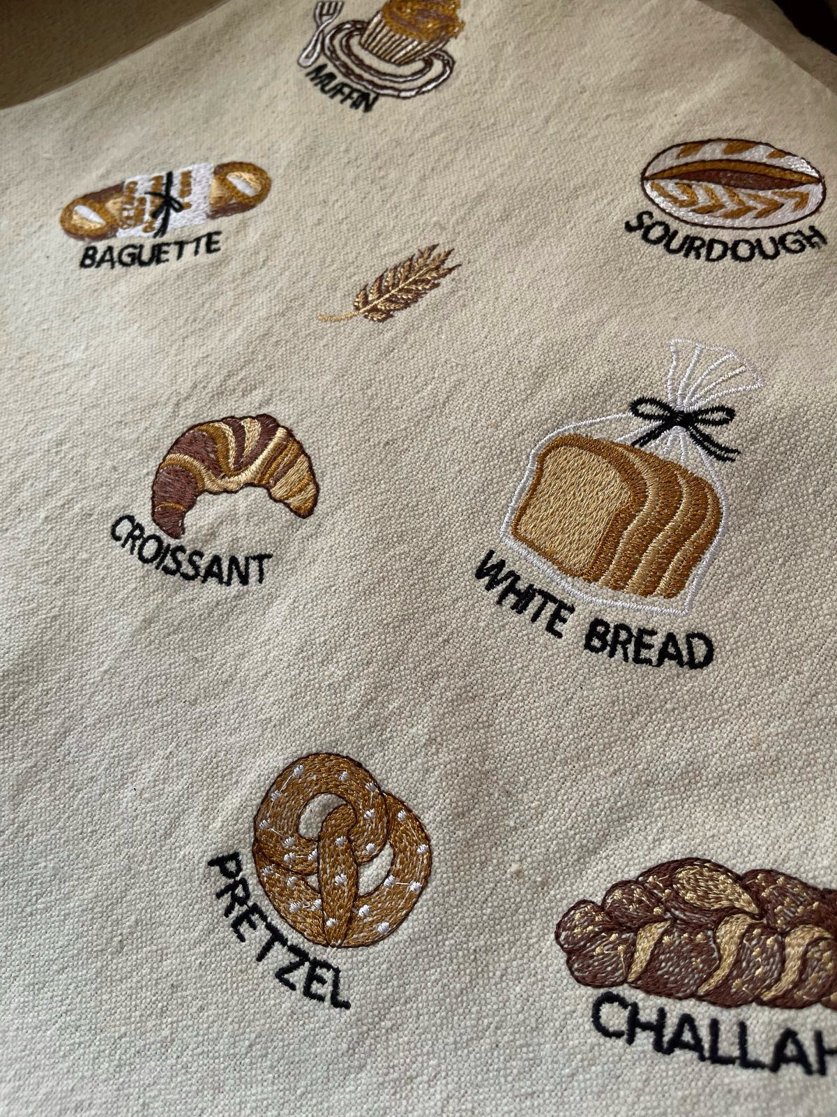 Beige tea towel with embroidered bread types and their names in black text.
